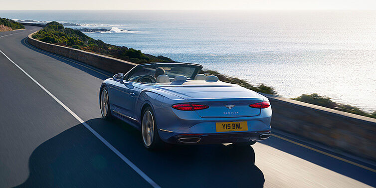 Bentley Praha New Bentley Continental GTC Azure convertible rear view in Light Windsor blue paint with 22 inch ten spoke wheels driving dynamically on an ocean road in the sunshine